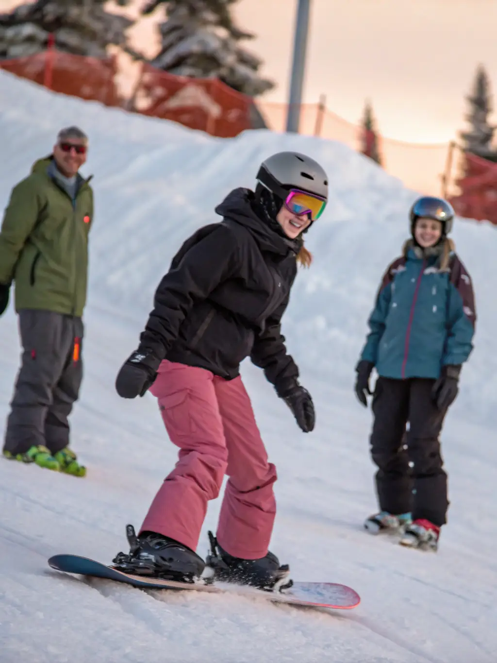 A young child in full ski gear, smiling and being assisted by a ski instructor on a gentle, snow-covered slope. The instructor is pointing towards the direction of travel, and the child looks eager and focused.